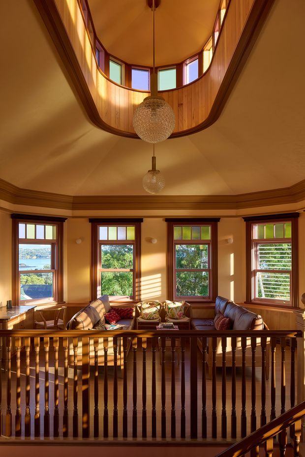 A richly detailed upper-level sitting area or landing in a historic-style home, enclosed by a curved timber balustrade and surrounded by tall timber-framed bay windows. Warm wood wall panelling, dark timber trim, decorative leadlight-style window panes, patterned sofas, and spherical pendant lights create an ornate, welcoming interior. Water and landscape are visible through the windows but are secondary to the interior architecture.