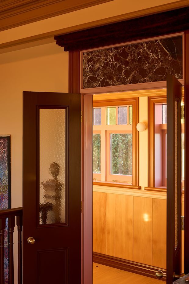A warmly lit traditional interior doorway with dark stained timber doors, textured glass panels, ornate timber trim, and a decorative marble transom panel. The open doorway reveals a wood-panelled room or hall beyond with timber-framed windows and polished timber flooring. Classical mouldings and rich amber lighting create an elegant heritage character.