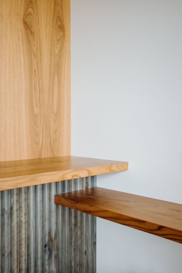 Close-up architectural detail of a contemporary timber stair or stepped landing. Thick natural timber treads and a timber wall panel are paired with a vertically ribbed, weathered metal-clad riser or wall surface against a plain light wall. The image emphasizes craftsmanship, grain, layered geometry, and contrasting warm wood with cool corrugated metal.