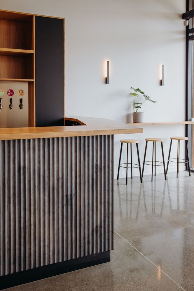A refined contemporary bar or hospitality-style interior featuring a large island counter with a light timber benchtop and vertically fluted dark ribbed frontage. Three slim black bar stools line a pale timber ledge beneath minimalist wall sconces. Built-in dark timber shelving and cabinetry occupy the left side, while polished concrete flooring, white walls, and extensive glazing create a clean, understated atmosphere.