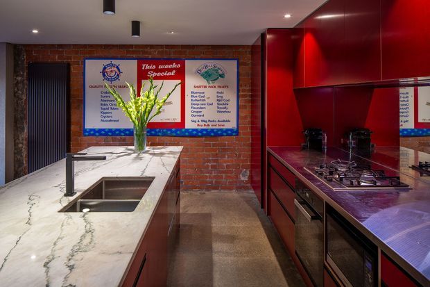 A compact commercial-style kitchen with a long white veined stone island and integrated double sink, dark red glossy cabinetry, gas cooktop, stainless appliances, exposed red-brick walls, polished concrete flooring, and menu-style wall signage. Reflective cabinetry creates mirror-like views of the cooking area. The space has a bold, industrial-inspired fit-out with dramatic red joinery and warm lighting.