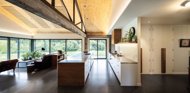 Open-plan contemporary kitchen and living area with a large central island, white built-in cabinetry, dark flooring, timber-lined ceiling and feature wall panels, exposed concrete and steel structural elements, and expansive glazing overlooking greenery. A mezzanine-like upper walkway or void is visible above the living space.