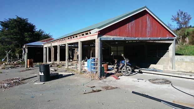 Exterior view of a large utilitarian workshop or shed with an open-sided concrete-framed lean-to extension. The main gabled structure has red corrugated metal cladding and a pale grey metal roof, while the foreground is a rough paved work yard with scattered construction materials and equipment. The building appears industrial or agricultural in character.
