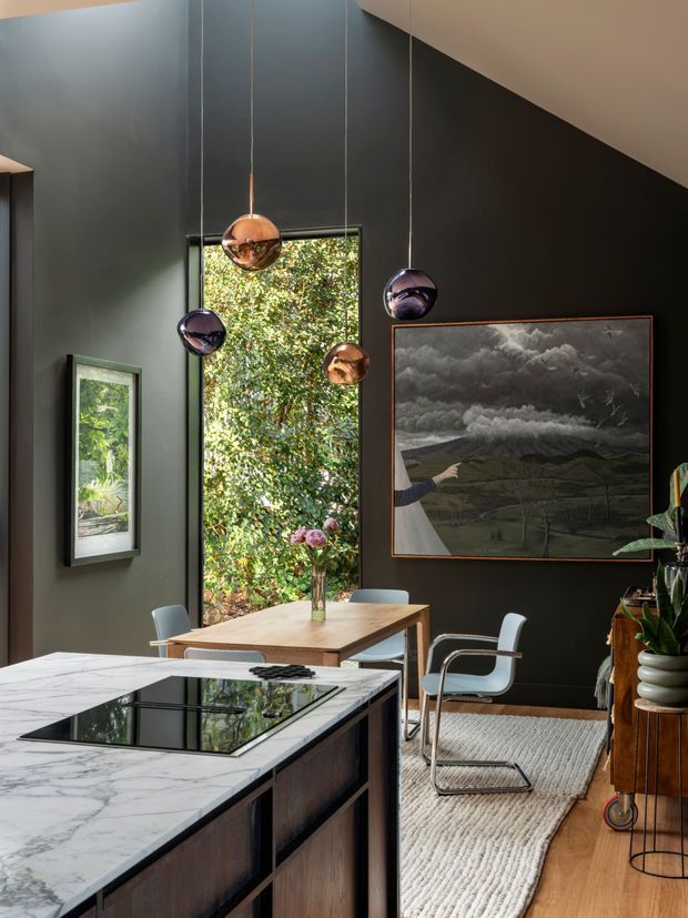 A sophisticated open-plan kitchen and dining area with a large white marble island in the foreground, dark wood cabinetry, charcoal walls, and a warm timber dining table. Sculptural pendant lights hang at varied heights beside a tall window overlooking lush greenery. A large dramatic landscape artwork, contemporary dining chairs, textured rug, timber flooring, and indoor plants create a moody yet refined interior.