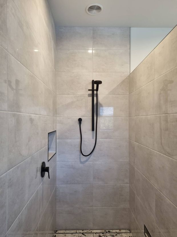Compact walk-in shower enclosure with large light grey stone-look wall tiles, matching tiled floor, a matte black handheld shower rail and mixer, recessed shelf, and ceiling exhaust vent. The space is clean, modern, and minimally detailed.