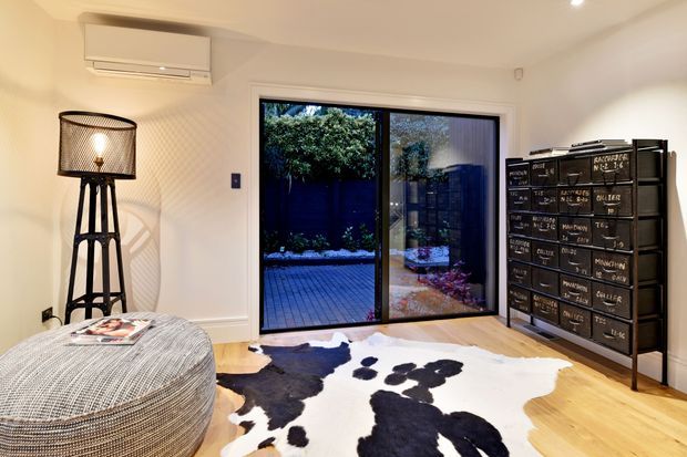 Bright contemporary living room with pale timber flooring, white walls, a large black-framed sliding glass door opening to a private courtyard, a sculptural black floor lamp, textured round ottoman, and distinctive dark multi-drawer storage cabinet. A black-and-white cowhide rug adds contrast.