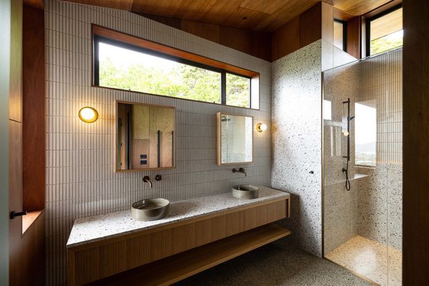 A warm, highly crafted bathroom with a double vanity, two round vessel basins, an open timber-fronted vanity, and a walk-in glass shower. Fine vertically ribbed wall tiles surround the room, while a terrazzo-like stone wall and benchtop add texture. Timber-clad walls and ceiling, bronze-toned window and door framing, circular wall sconces, and generous horizontal glazing create a mid-century-inspired, nature-connected interior.