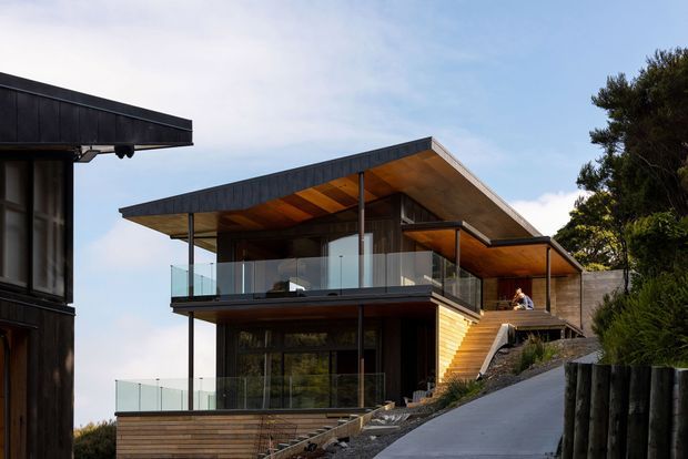 Exterior view of a contemporary multi-level house on a sloped site, featuring broad asymmetrical pitched roof planes, extensive floor-to-ceiling glazing, dark cladding, warm timber soffits and walls, and glass balcony balustrades. A driveway rises toward the house, with surrounding trees and neighbouring building elements visible at the edges.