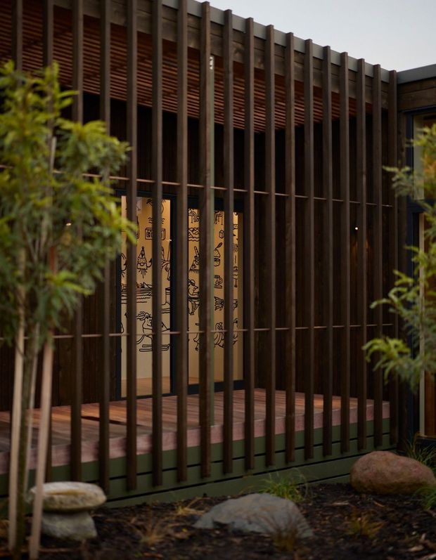 A close-up exterior view of a contemporary building facade screened by tall, closely spaced dark timber or metal vertical fins. Warm timber soffit slats and a timber deck frame a softly lit glazed entry or interior area behind the screen, with illustrated panels visible inside. Landscaping with shrubs, rocks, and mulch occupies the foreground.