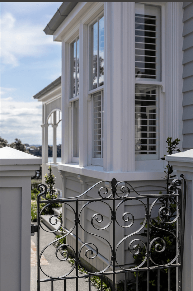 Close-up exterior view of a refined light-coloured rendered and weatherboard-clad building, featuring tall multi-pane windows with horizontal security bars, substantial decorative white window surrounds, and an ornate black wrought-iron gate. A covered architectural walkway or veranda with classical arched detailing is visible in the background.