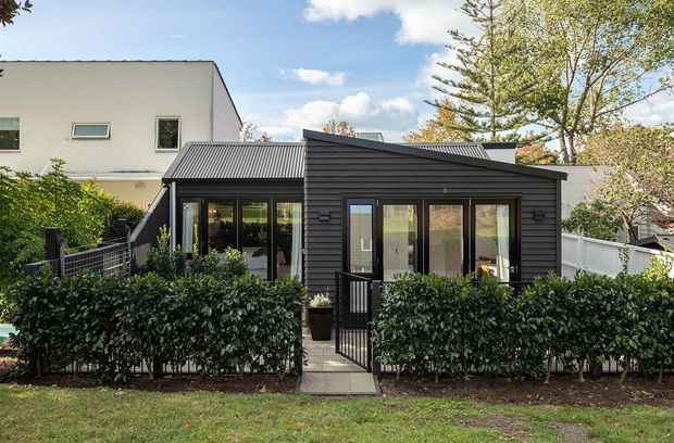 Exterior view of a compact contemporary single-storey house or extension with dark horizontal cladding, a low pitched metal roof, and large glazed doors and windows. A small gated path leads through a hedge-lined front garden to the entry. A taller light-coloured neighbouring building and mature trees are visible in the background.