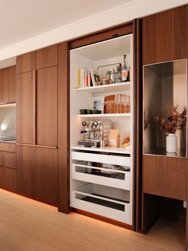 Close-up view of a sophisticated built-in timber wall with an open bar and coffee station. The dark walnut-toned cabinetry incorporates open white shelving, multiple drawers, a coffee machine, glassware, bottles, books and decorative objects. A recessed display niche with a vase is visible to the right, while warm integrated floor-level lighting and pale timber flooring create a refined, inviting atmosphere.