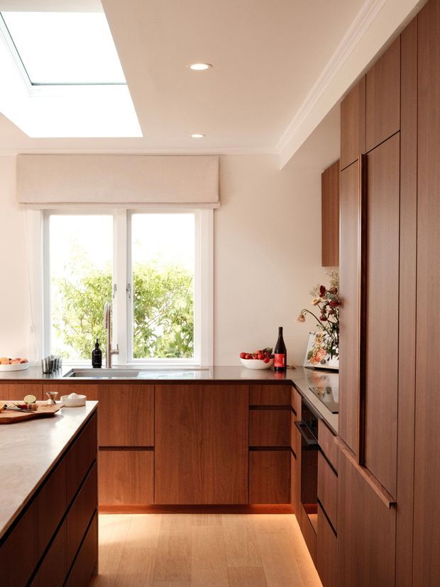 Warm, refined contemporary kitchen with extensive dark timber cabinetry, a central island, dark stone benchtops, integrated appliances, a large window, and a skylight. The space is softly lit with recessed ceiling lights and under-cabinet illumination, creating a welcoming, polished atmosphere.