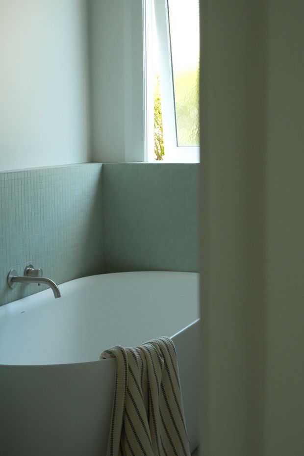 A quiet, minimalist bathroom corner featuring a freestanding white bathtub beneath a narrow frosted-glass window. Small pale blue-green mosaic tiles line the lower walls, while smooth light walls and a brushed metal wall-mounted faucet create a clean, softly lit atmosphere. A striped neutral towel hangs over the bath edge.