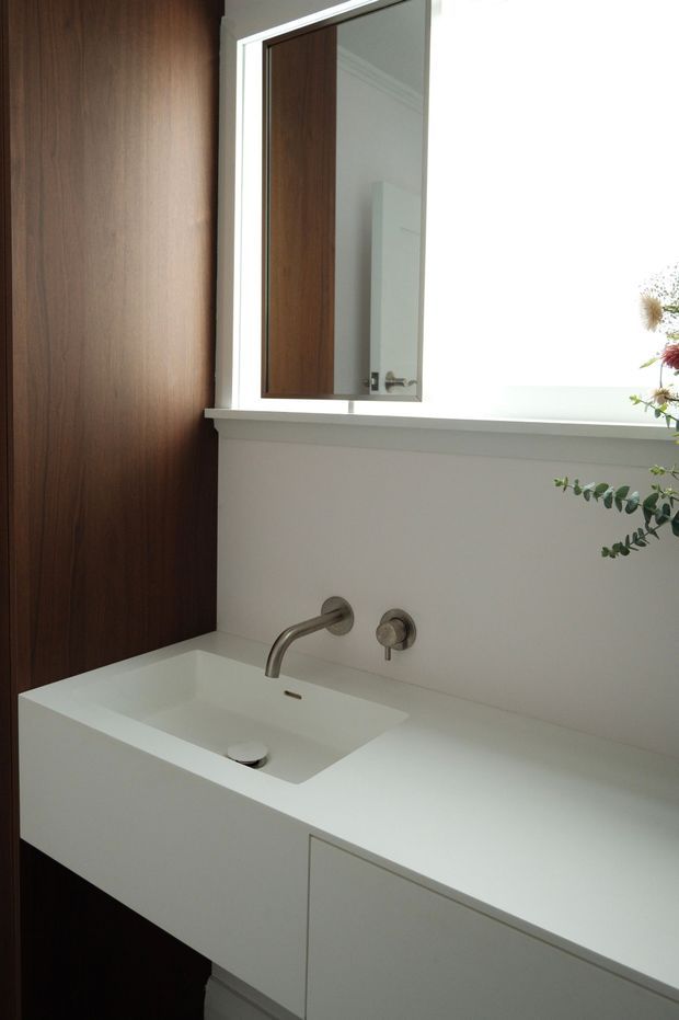 A bright, minimalist bathroom vanity with a white integrated rectangular basin and wall-mounted brushed metal tapware. The vanity has clean-lined white cabinetry and a broad white benchtop, set against a dark timber wall panel. A large mirrored cabinet or mirror is mounted above the basin, with a bright window and a small floral arrangement at the right edge.