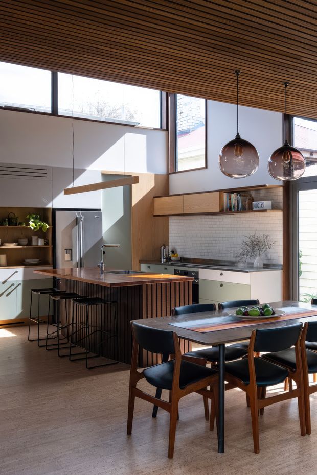 A bright, open-plan kitchen and dining area with a warm timber slatted ceiling, full-height and clerestory glazing, pale walls, and timber cabinetry. The kitchen features a long island with dark vertical slatted detailing, a stainless-steel sink and tap, integrated appliances, pale green and white cabinetry, and a small white tiled splashback. A dark timber dining table with mid-century-style upholstered timber chairs occupies the foreground, while two large smoky glass pendant lights hang above. Natural light creates strong geometric shadows across the room.