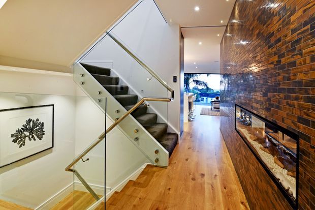 A polished residential hallway with a timber floor, a glass-and-metal balustraded staircase, recessed ceiling lights, and a dramatic full-height dark brick feature wall incorporating a long linear fireplace. The corridor opens toward a brighter living area and exterior glazing in the distance.
