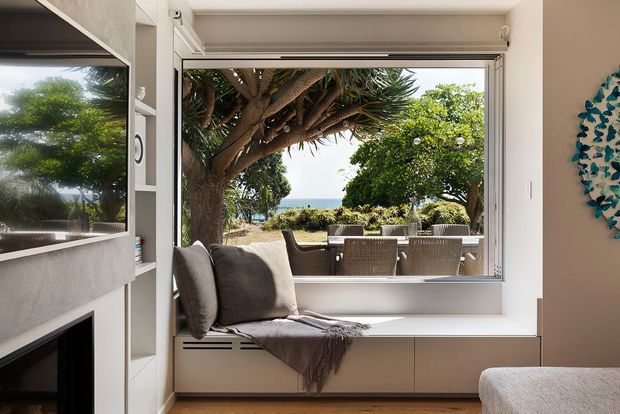 A bright contemporary living room with a built-in window seat overlooking a landscaped outdoor terrace and garden. The room features a large wall-mounted television, a black fireplace, white built-in shelving and cabinetry, a deep-set picture window with a cushioned bench, and neutral upholstered seating. The open window reveals outdoor dining furniture and lush planting beyond.