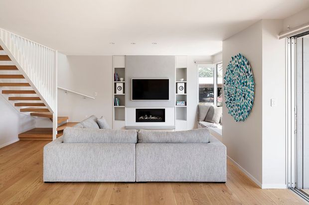 Bright contemporary living room viewed from behind a large light-grey sectional sofa. The room features warm timber flooring, white walls and ceiling, a built-in white media wall with open shelving and fireplace, and an open-riser timber staircase with white balustrade. A glazed opening and vertical blinds are visible at right, with a blue patterned wall artwork adding colour.