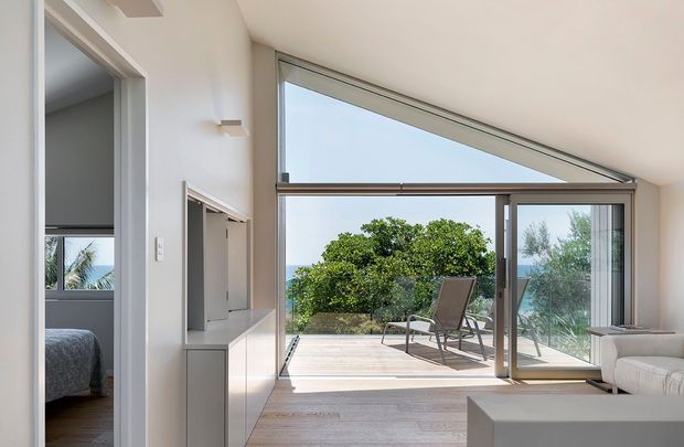 Bright contemporary interior opening onto a timber deck and leafy coastal outlook. The room features a large angular floor-to-ceiling glazed wall with sliding glass doors, pale timber flooring, white walls and ceiling, and minimal built-in cabinetry. A bedroom is visible through the doorway on the left, while outdoor seating and vegetation are visible beyond the glazing.