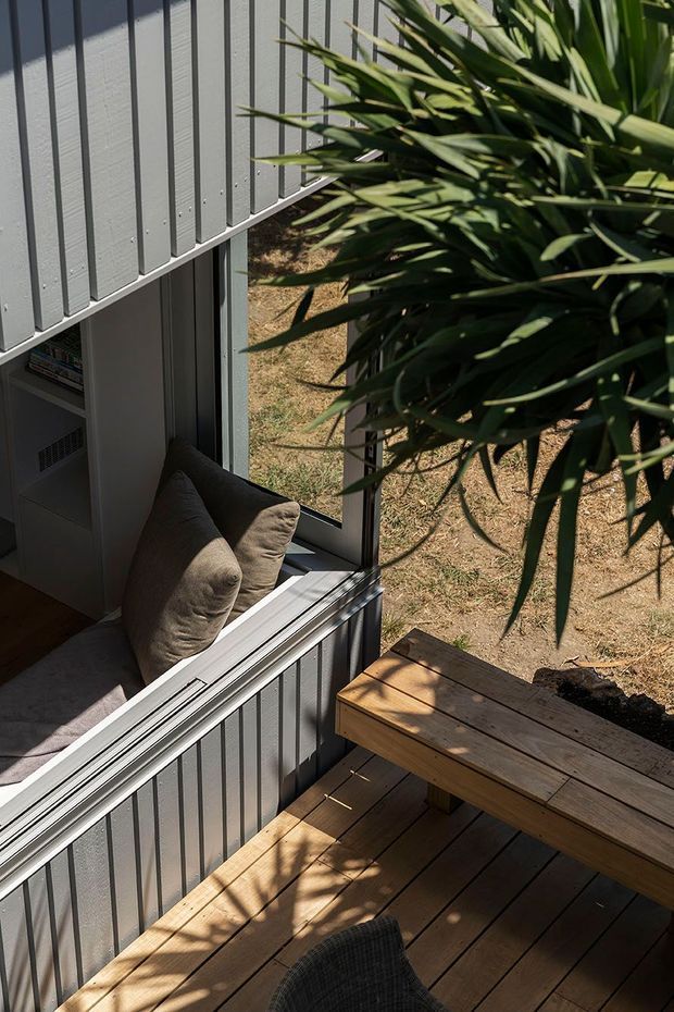 A compact indoor-outdoor seating area viewed from above, with a timber deck and built-in timber bench/table beside a large opening. The interior includes a cushioned window seat, vertical grey wall cladding, and dark-framed glazing, while leafy planting partially screens the deck. Strong sunlight creates a warm, casual atmosphere.