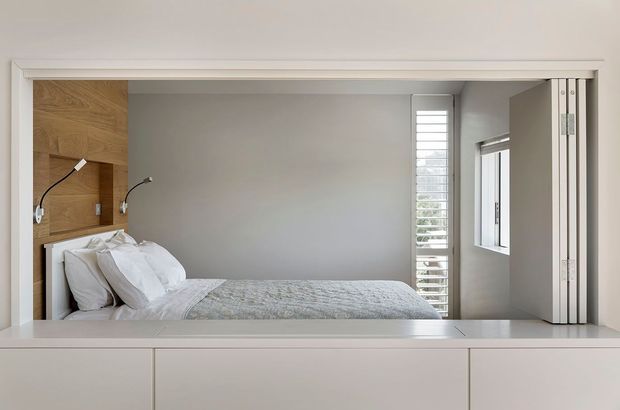 A compact, light-filled bedroom viewed through a wide internal opening or pass-through. The room contains a bed with white bedding, a timber feature wall behind the headboard with two wall-mounted reading lights, white built-in cabinetry below the opening, and slim white-framed windows with louvred screening. The design is clean and restrained with a neutral palette and modern built-in detailing.
