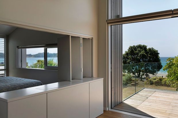 A bright contemporary interior with a built-in white storage partition separating a bedroom from an adjacent living area. The bedroom is partially visible through the open partition, while full-height glazed doors open onto a timber deck with a frameless glass balustrade and views toward the water. Clean-lined joinery, pale walls, timber flooring, and abundant natural light create an airy coastal setting.