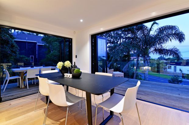 Bright contemporary dining area with a large dark dining table and white molded chairs, opening through expansive black-framed glazing onto a timber deck and landscaped outdoor setting. The room has warm timber flooring, white walls and ceiling, and strong indoor-outdoor connection.