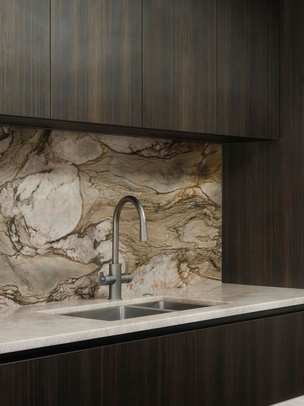 Close-up view of a refined kitchen sink area with dark vertically grained cabinetry, a large dramatic veined natural-stone splashback and matching benchtop, and a brushed metal gooseneck tap over an undermount sink. The palette is dark, warm, and luxurious with strong natural stone patterning.