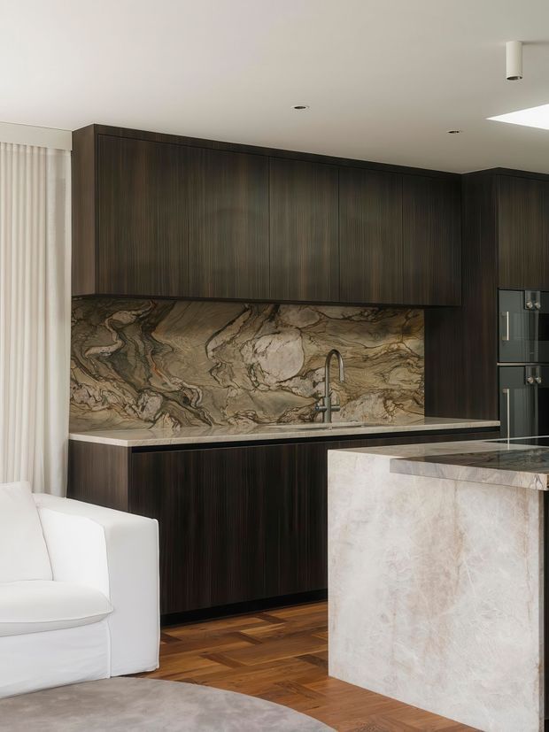 A refined open-plan kitchen with dark vertically grained timber cabinetry, a dramatic veined natural-stone splashback and matching island cladding, light stone benchtops, integrated dark appliances, warm timber flooring, and a white upholstered sofa at the edge of the frame. The space has a restrained, luxurious contemporary appearance with recessed and understated ceiling lighting.