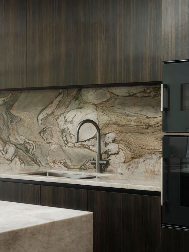 Close-up view of a sophisticated contemporary kitchen featuring dark vertically grained timber cabinetry, a dramatic veined natural stone splashback and benchtop, an undermount sink with a curved dark tap, and integrated black appliances. A stone island edge is visible in the foreground.