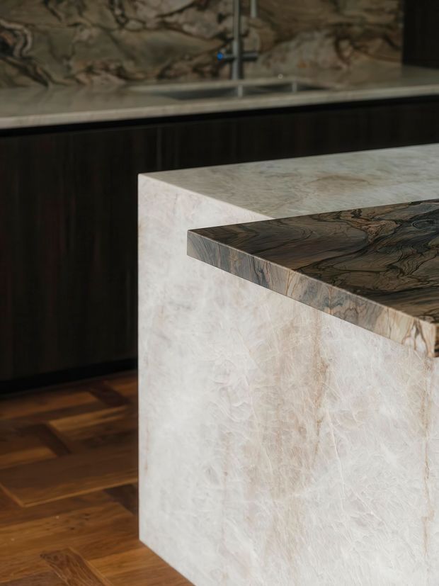 Close-up view of a luxurious kitchen island with a pale veined stone waterfall-style base and benchtop, paired with a dark, highly figured stone overhang. Dark timber cabinetry, a reflective stone splashback or wall surface, and warm timber flooring are visible in the background.