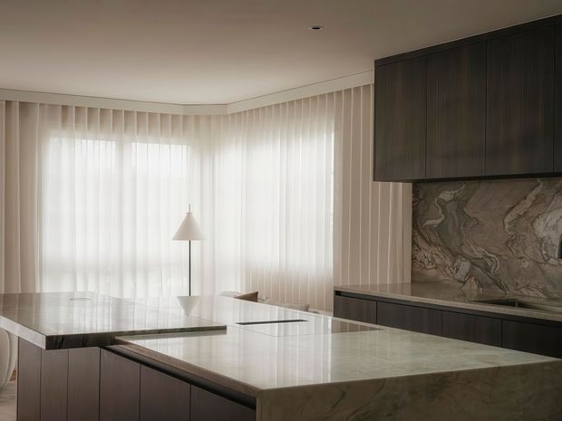 A refined contemporary kitchen with a large waterfall-edge stone island and matching stone benchtops. Dark timber-grain cabinetry contrasts with a dramatic veined stone splashback, while sheer floor-to-ceiling curtains provide soft natural light. A sculptural pendant light and minimal detailing create a calm, luxurious atmosphere.