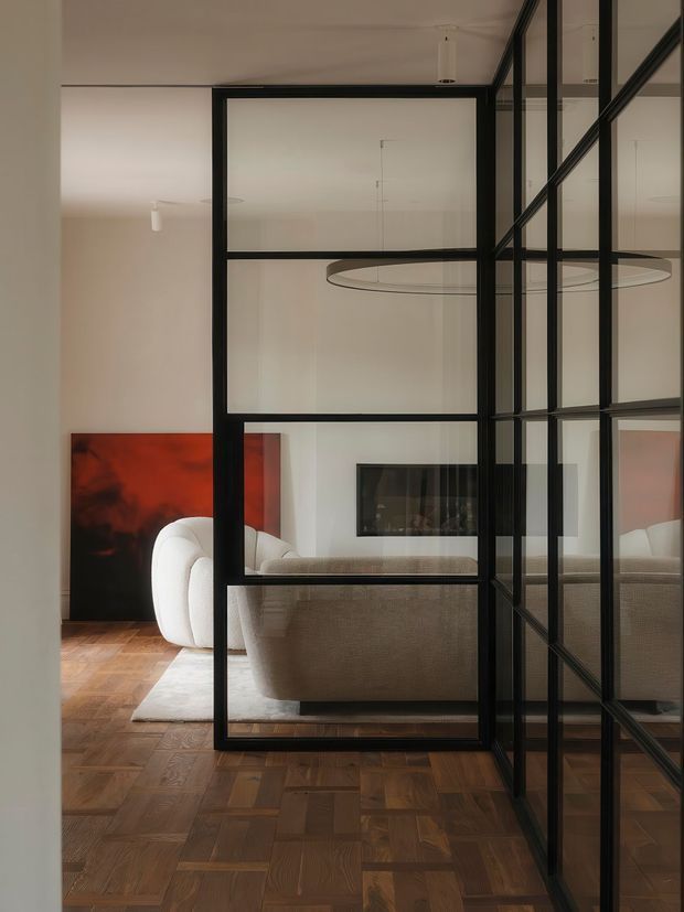 Interior view through black metal-framed glass partitions into a refined living room. The space features warm parquet timber flooring, pale walls, a cream sculptural sofa, a neutral upholstered sofa, a large circular pendant light, and a bold red-and-black artwork. Reflective glazing and strong black grid framing create a modern, graphic composition.