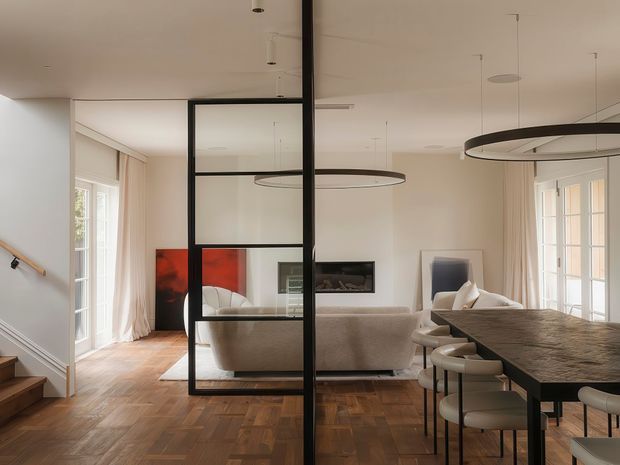 Open-plan contemporary living and dining area viewed from an entry/stair zone. The space features warm parquet timber flooring, pale walls, large glazed doors and windows with sheer curtains, a neutral upholstered sofa, a dark stone-look dining table with upholstered stools, and a black-framed glazed partition dividing the circulation area from the living room. Sculptural circular pendant lights and artwork add restrained modern detailing.