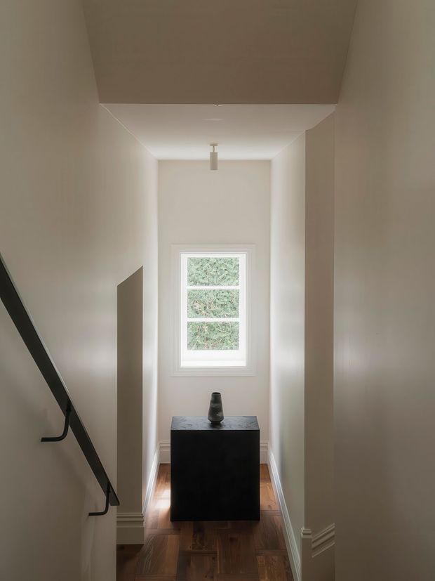 A narrow, softly lit hallway or stair landing with warm painted walls, white trim, and a timber floor. A black floating stair handrail runs along the left wall, while a dark console cabinet with a small vase sits beneath a tall multi-pane window at the end. The space has a restrained, contemporary and minimalist appearance with gentle natural light.