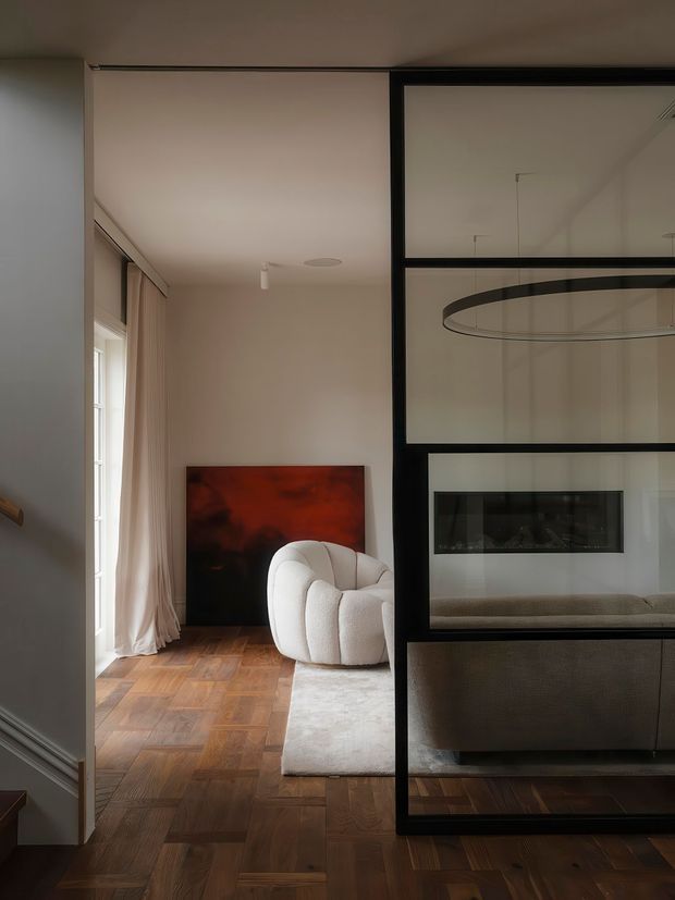 Refined contemporary living room with warm parquet flooring, soft neutral walls, floor-length curtains, a sculptural cream lounge chair and a large abstract artwork. A black-framed glazed partition separates the seating area from a fireplace zone, with a large circular pendant light visible through the glass. The space has a calm, sophisticated, moderately dim atmosphere.