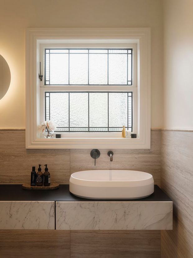 A refined bathroom vanity with a white vessel basin on a dark benchtop, integrated wall-mounted tapware, marble-look drawer fronts, and warm wood-look wall tiling. A large frosted leadlight-style window with layered white trim dominates the wall above the vanity, complemented by a round wall light and small toiletries.