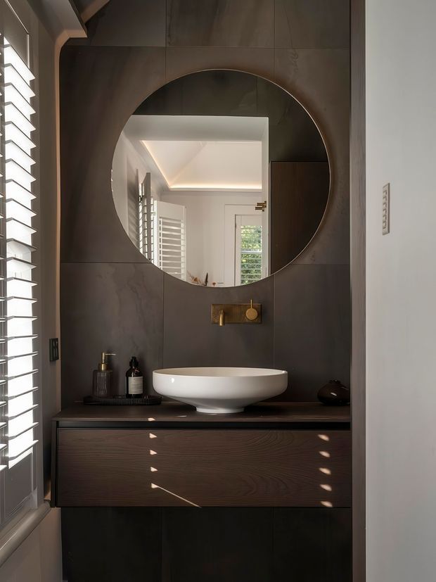 A compact, refined bathroom vanity area featuring a round wall mirror set into dark stone-look wall panels, a white vessel basin on a dark benchtop, brushed brass wall-mounted tapware, and dark timber-grain floating cabinetry. Plantation shutters on the side window cast striped sunlight across the joinery and wall.