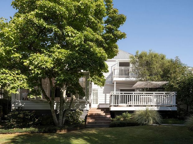 A sunny exterior view of a white weatherboard multi-level home, partially screened by mature leafy trees. The house features a pitched corrugated roof, upper and lower timber verandah-style decks with white balustrades, timber stairs, and a landscaped lawn with ornamental grasses.