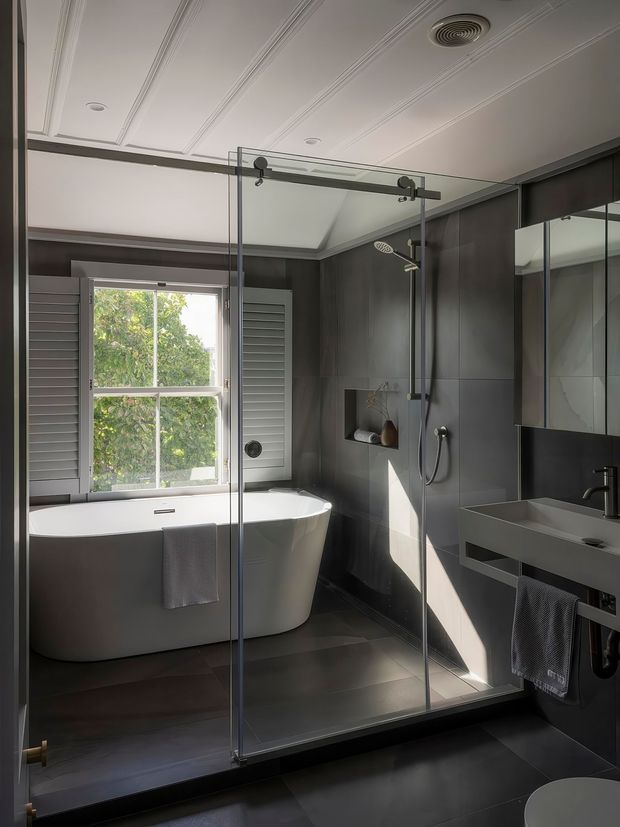 A sophisticated contemporary bathroom with a freestanding white bathtub beside a shuttered window, a frameless glass walk-in shower, dark grey wall and floor finishes, and a floating grey vanity with integrated basin and black tapware. White decorative ceiling panelling and strong natural light create contrast against the dark surfaces.