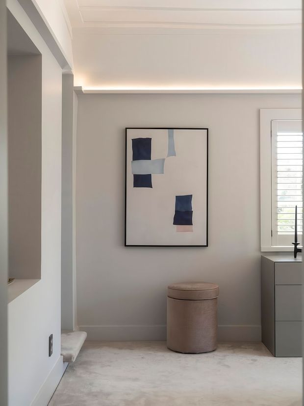 A refined, softly lit interior with pale grey walls, white decorative ceiling detailing and concealed cove lighting. A framed abstract artwork hangs above a round upholstered ottoman, with built-in cabinetry and a shuttered window on the right. The room has a calm, minimalist character and appears to be a compact dressing or wardrobe area.