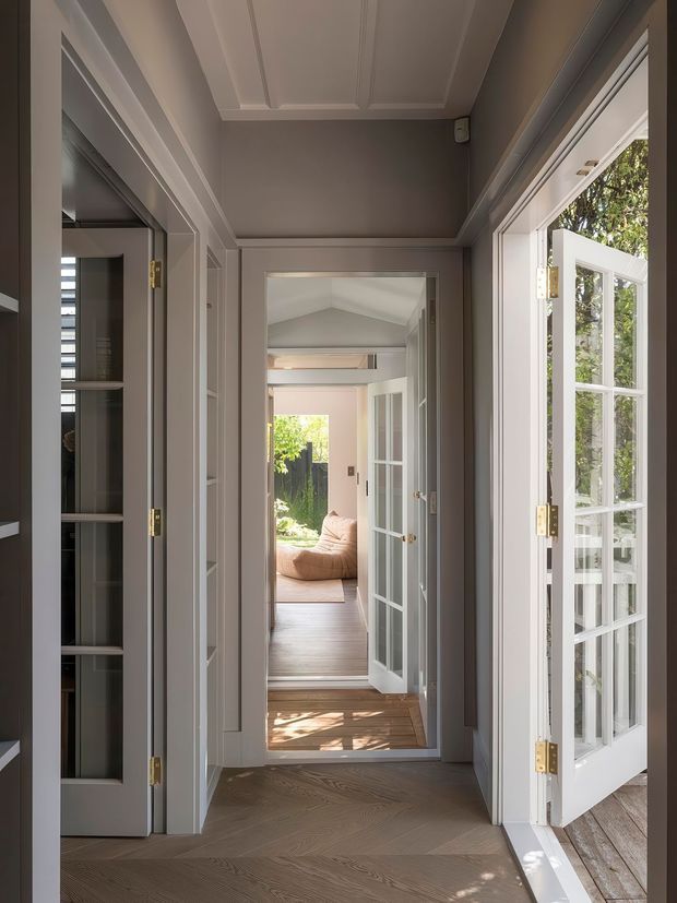 A bright, refined interior hallway viewed through a series of open glazed doors, with dark timber herringbone flooring, pale grey-beige walls, white panelled trim, and a coffered ceiling. The corridor leads toward a sunlit living area with an upholstered sofa visible in the distance. Tall French doors on the right open to an exterior deck or garden, bringing in natural light.