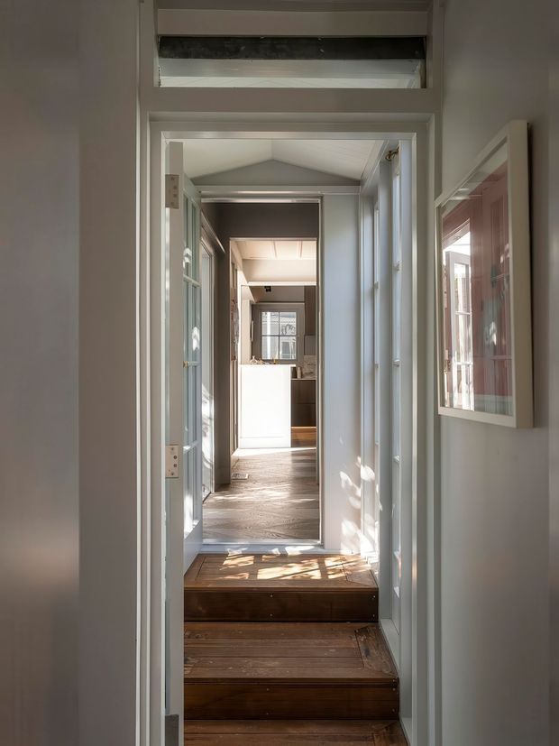 A narrow, sunlit interior passage with several timber steps leading through white-painted doorways toward a bright kitchen area. The corridor features white trim, glazed internal doors, a pitched ceiling, timber flooring and stair treads, and framed wall artwork. Strong natural light creates patterned shadows across the steps and floor.