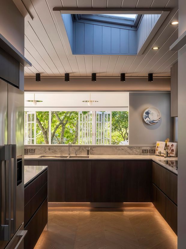 Contemporary kitchen with dark wood lower cabinetry, pale stone benchtops, integrated appliances, and a large window overlooking leafy greenery. The room features a white paneled ceiling with recessed spotlights, a blue-lined skylight, under-cabinet lighting, and a compact island or peninsula. A decorative plate is mounted on the grey wall beside the window.