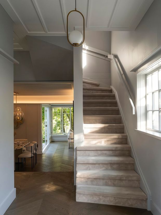 A refined residential stairway and landing with a broad carpeted staircase, white-painted walls and ceiling with decorative recessed panelling, and a large multi-pane window bringing in natural light. A sculptural brass-and-opal pendant hangs above the stair, while a dining area and bright glazed sitting space are visible beyond the hallway. The floor is laid in a dark herringbone timber pattern.