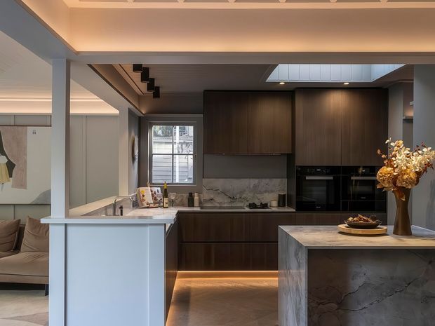A refined contemporary kitchen viewed from the adjoining living area, featuring dark timber cabinetry, a marble-look stone splashback and island, integrated black appliances, warm under-cabinet lighting, and a skylight above the cooking zone. A partial living room and staircase are visible beyond the kitchen.