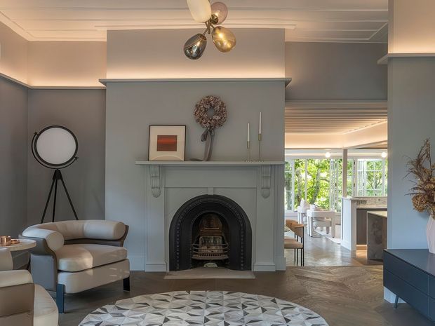 A refined living room centred on a decorative fireplace with a dark arched surround and pale grey mantel. The room features a sculptural cream sectional sofa, geometric patterned rug, dark built-in console, tripod floor lamp, framed artwork, and a polished multi-globe pendant light. Herringbone timber flooring, detailed ceiling moulding, and glazed openings connect the living area to a bright adjoining space.