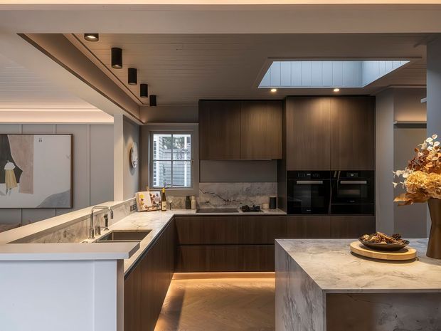 Contemporary kitchen with dark timber cabinetry, pale marble-look benchtops and splashback, integrated black appliances, under-cabinet and toe-kick lighting, and a large central island. The room features a paneled ceiling with recessed spotlights and a prominent skylight, with a dining or living area visible beyond the kitchen.