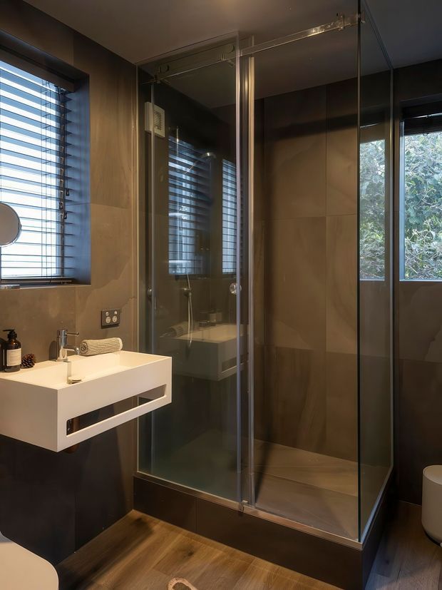 Compact contemporary bathroom with a frameless glass corner shower, floating white vanity, dark wall panels, warm wood-look flooring, and horizontal window blinds. The space has a moody, refined atmosphere with natural light and greenery visible through the window.